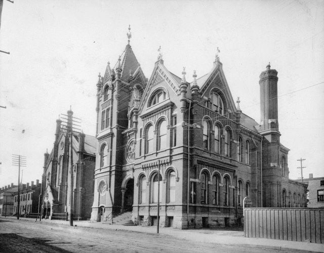 The first Hamilton Public Library, seen from the southeast from Main Street West, in a black & white photo. Centenary Methodist Church (now New Vision United Church) is seen to the west of the Victorian library building. The library is a neo-Romanesque building, with pointed roofs, round arches over the windows, and decorative brick details.