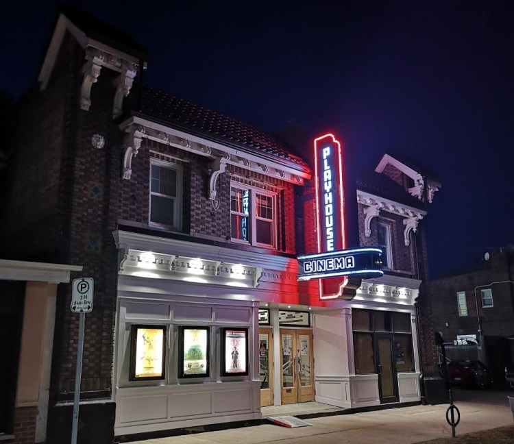 The Playhouse Cinema on Sherman Ave North, in Hamilton, Ontario, seen illuminated at night. It has a sloped roof, two-tone brickwork, white wooden trim and dentellation, white wooden panneling on the ground level, and a blue & red neon sign.