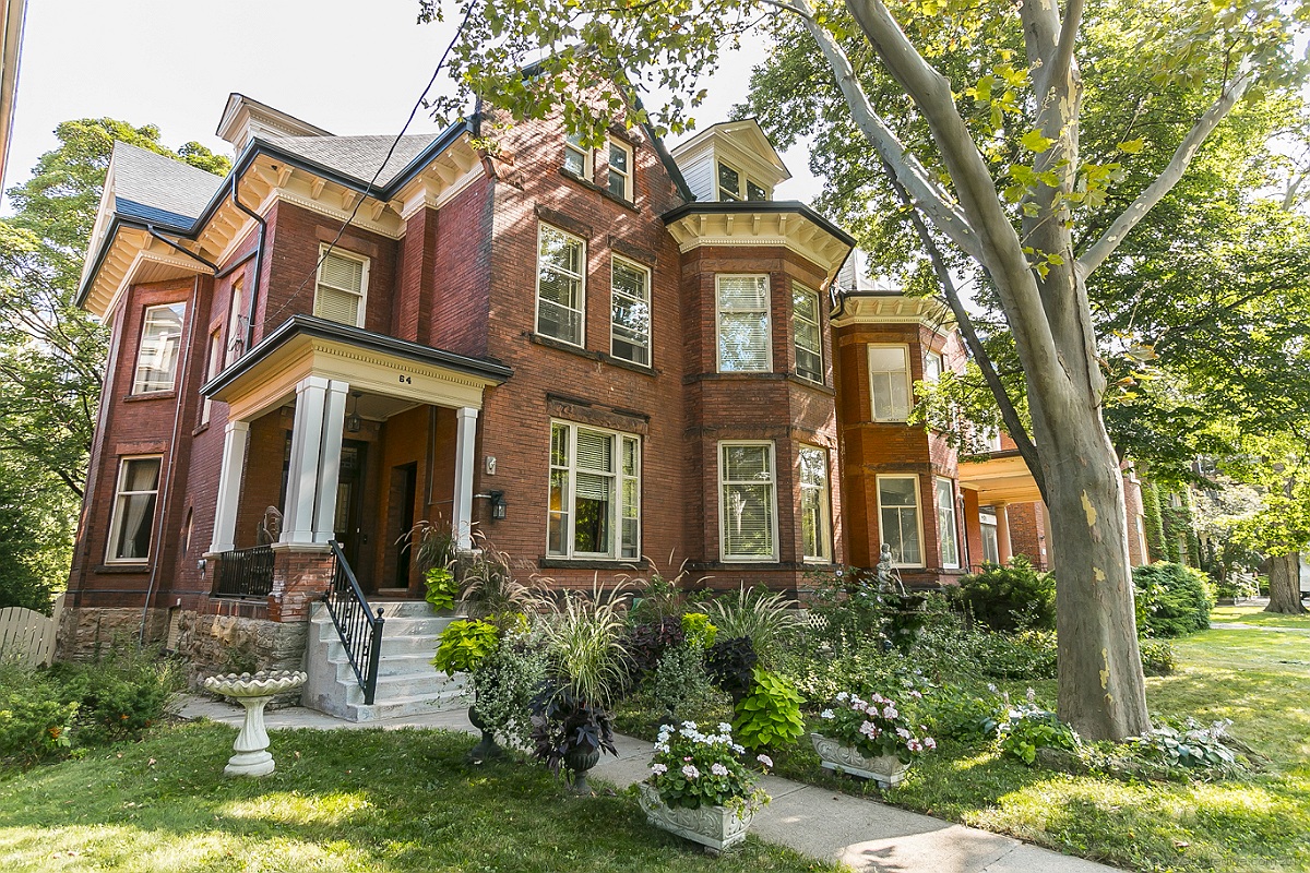 The westernmost of two grand, late-Victorian semis in red brick with cream trim and a tall old tree in the front, in Durand, Hamilton. Seen on a sunny day, with sunlight dappling the lush front garden.