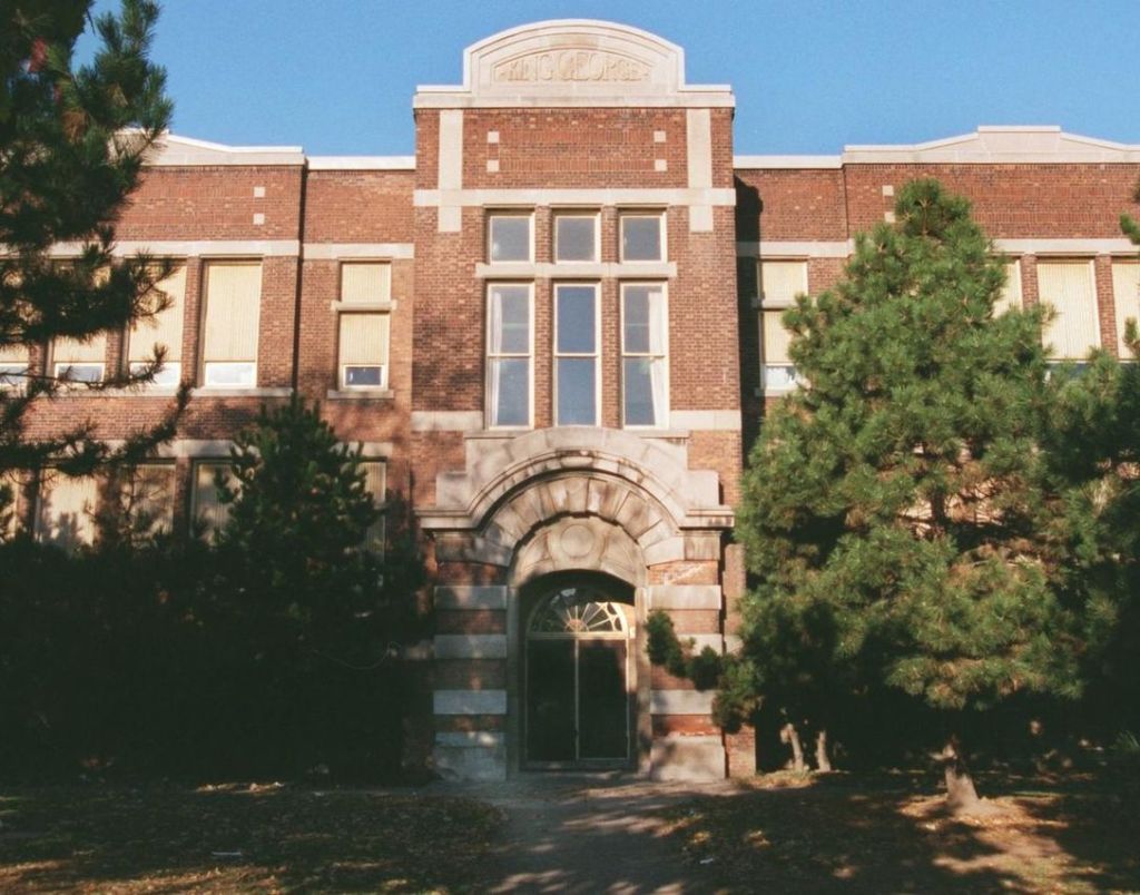 Dignified school building topped with name plaque reading KING GEORGE and grand entrance portico flanked by pine trees.