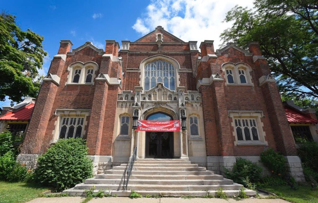 Gothic Revival church in brick and stone, flanked by mature trees, with red banner over the entrance advertising a contents auction with TimewellAuctions.com